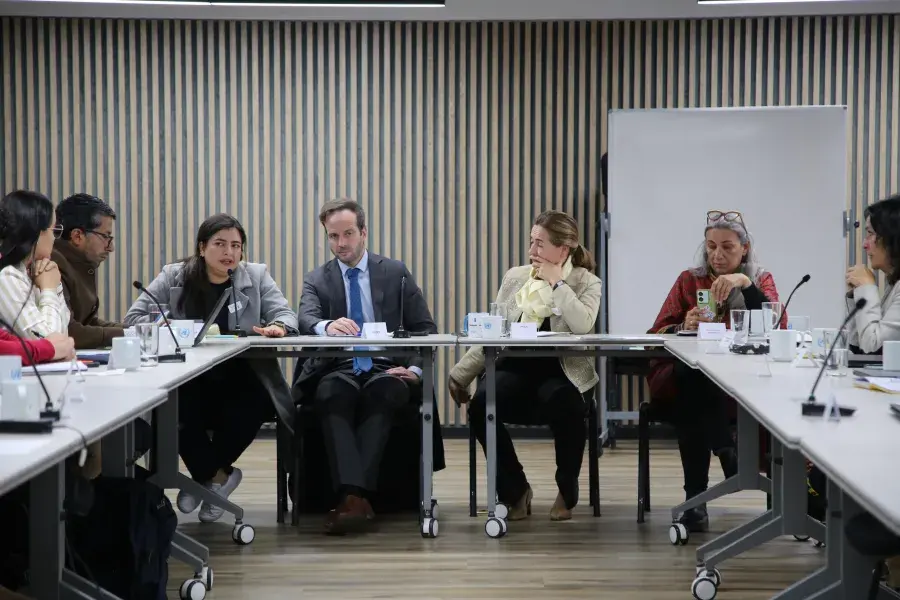 eunión de trabajo en una sala moderna con decoración minimalista y plantas. Ocho personas están sentadas alrededor de una mesa en forma de U, con laptops, tabletas, cuadernos y tazas. En el fondo, una pantalla muestra una presentación titulada "Fases del Proceso", con un cronograma de actividades entre abril y septiembre. Una mujer de pie, con mascarilla, sostiene un celular y observa la escena. Todos los participantes parecen estar atentos; algunos toman notas, otros escuchan o miran la pantalla.